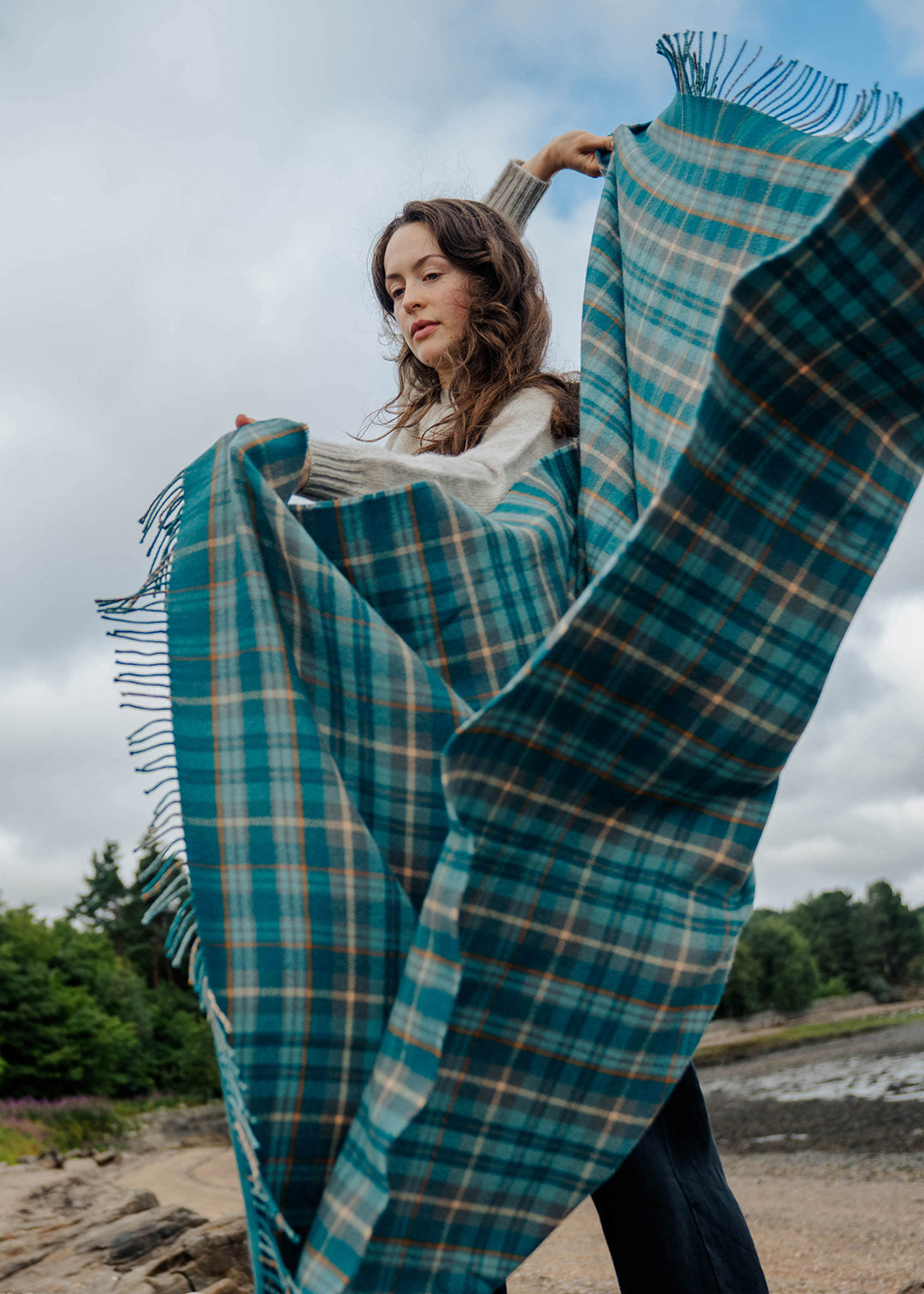 Model holding a lambswool teal tartan blanket on a beach.
