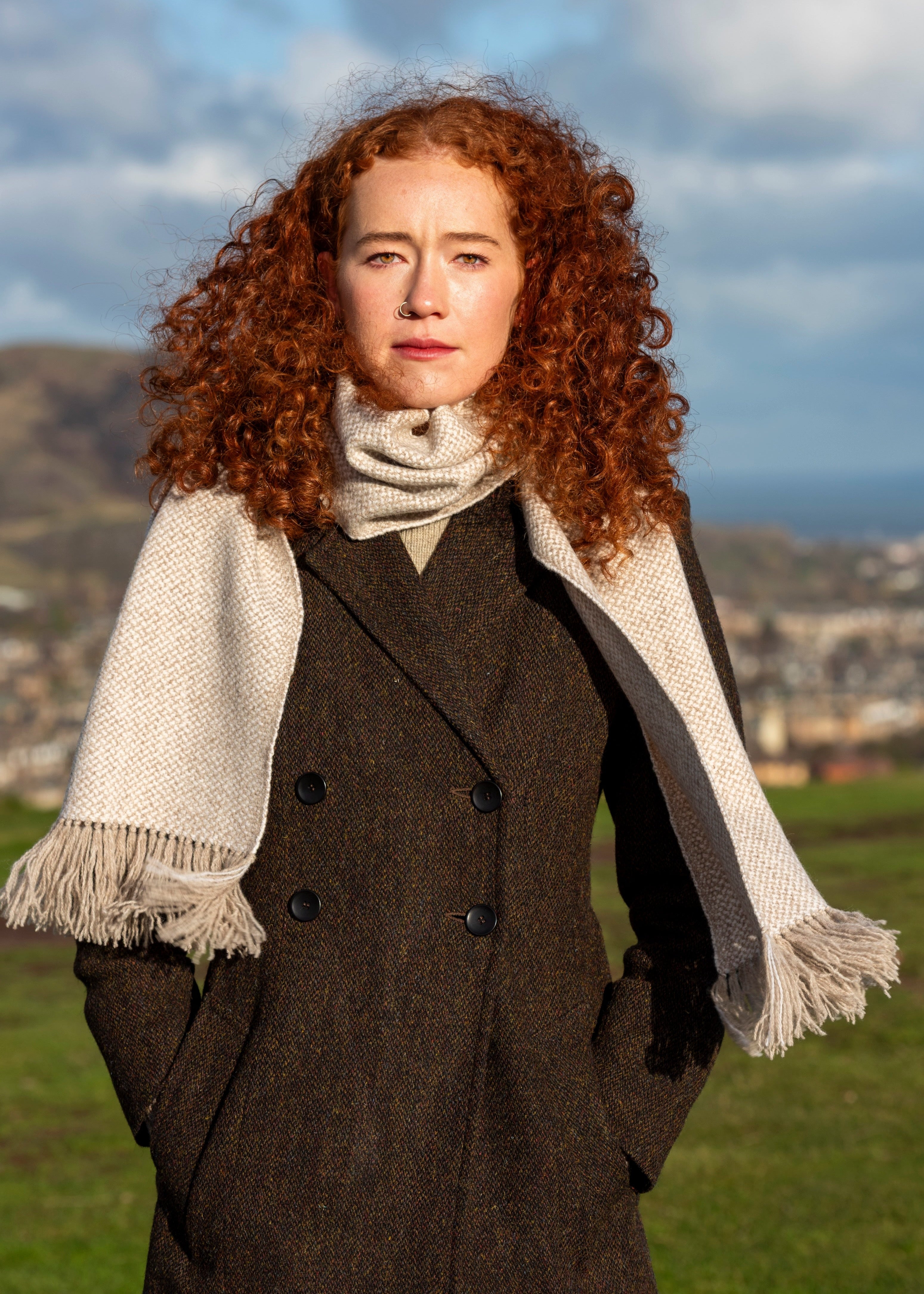 Woman with red hair wearing a brown Harris Tweed coat and hand woven british wool Glisk scarf in a scenic outdoor setting.