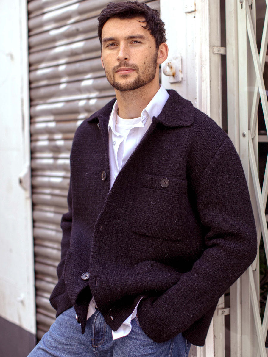 A man wearing a dark charcoal textured knit cardigan over a white collared shirt and blue jeans, leaning against a white metal shutter door.