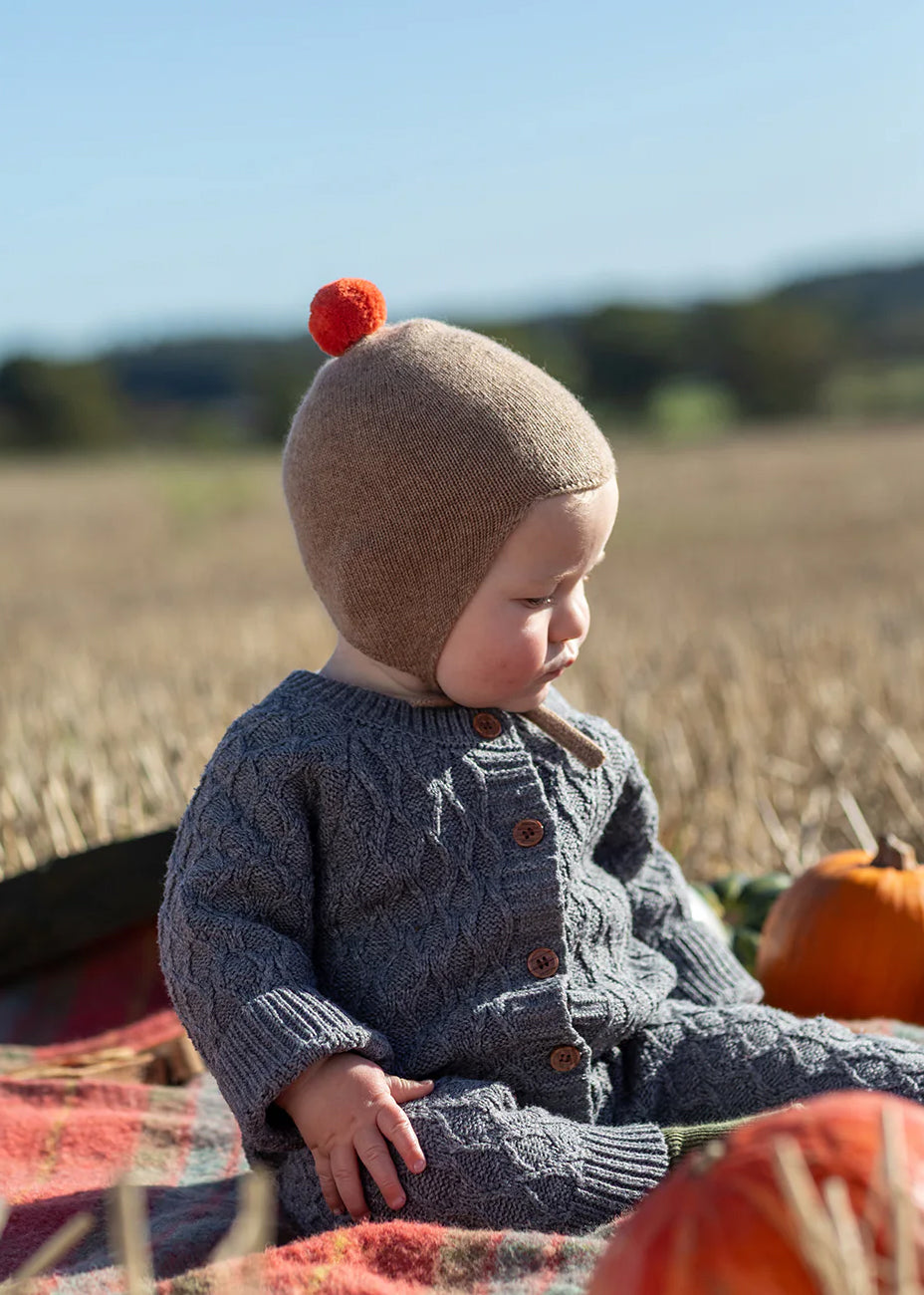 Cashmere baby bonnet in camel with a vibrant orange pom pom.