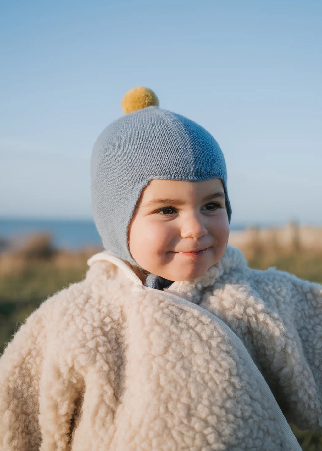 Cashmere baby bonnet in dusky blue with a yellow pom pom.