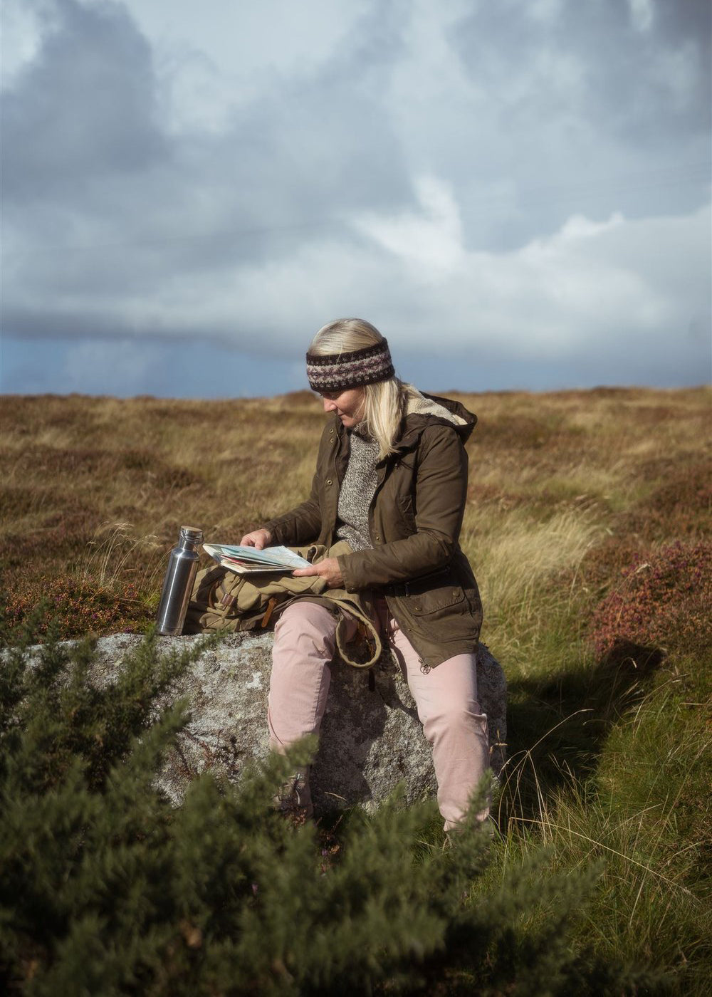 Myrtle knitted headband shown worn by Meg, the designer of the knitting pattern, sitting on a rock in the Scottish landscape.