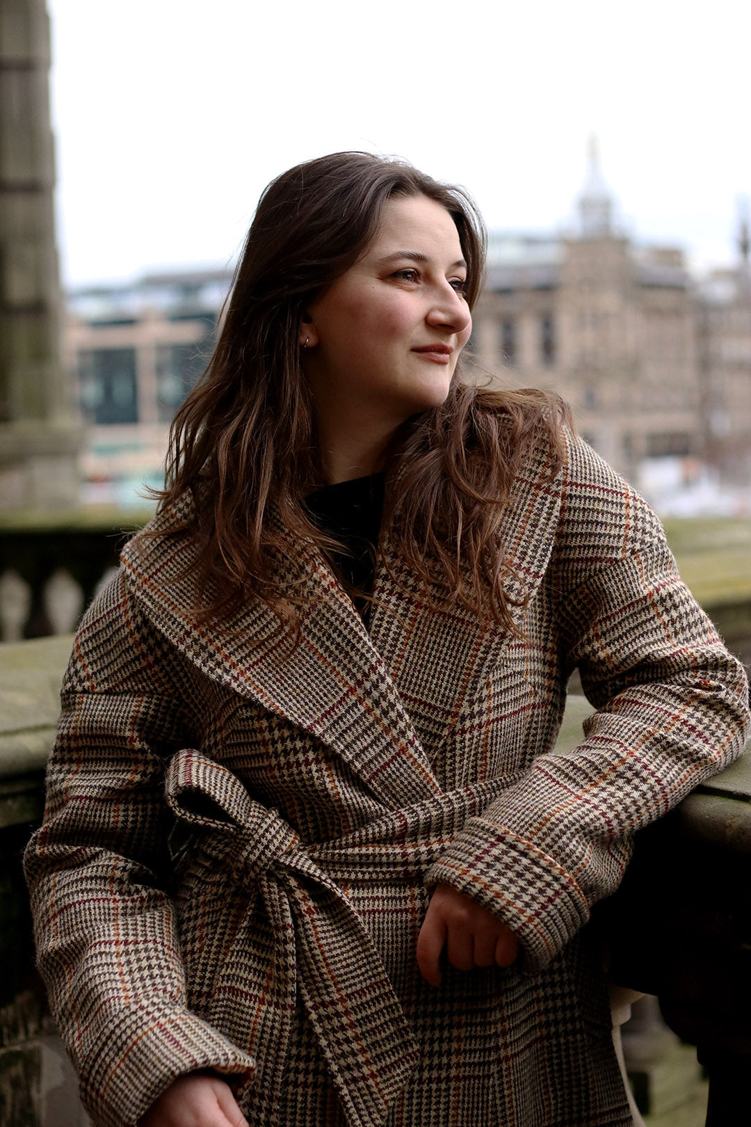 Harris Tweed checked Cora coat in camel with burgundy and ochre check, shown on model with blurred cityscape in background.