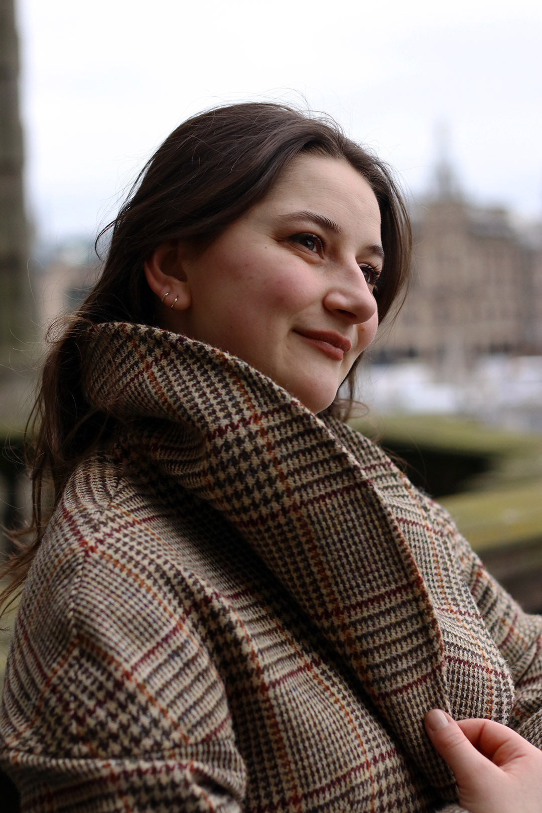 Harris Tweed checked Cora coat in camel with burgundy and ochre check, shown on model with blurred cityscape in background.