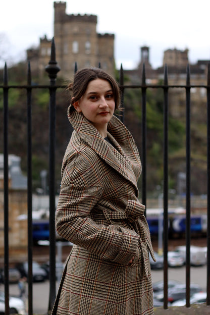 Harris Tweed checked Cora coat in camel with burgundy and ochre check, shown on model with blurred cityscape in background.