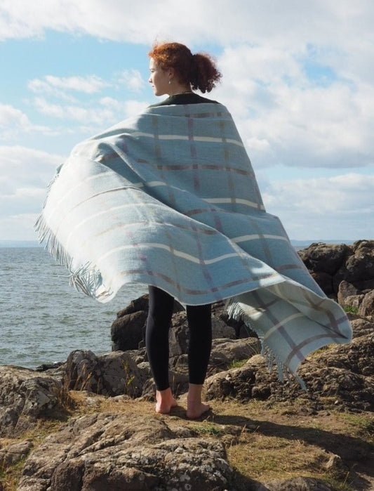 Maree throw woven in pure Merino Lambswool, wrapped around shoulders of model standing on rocky shoreline.