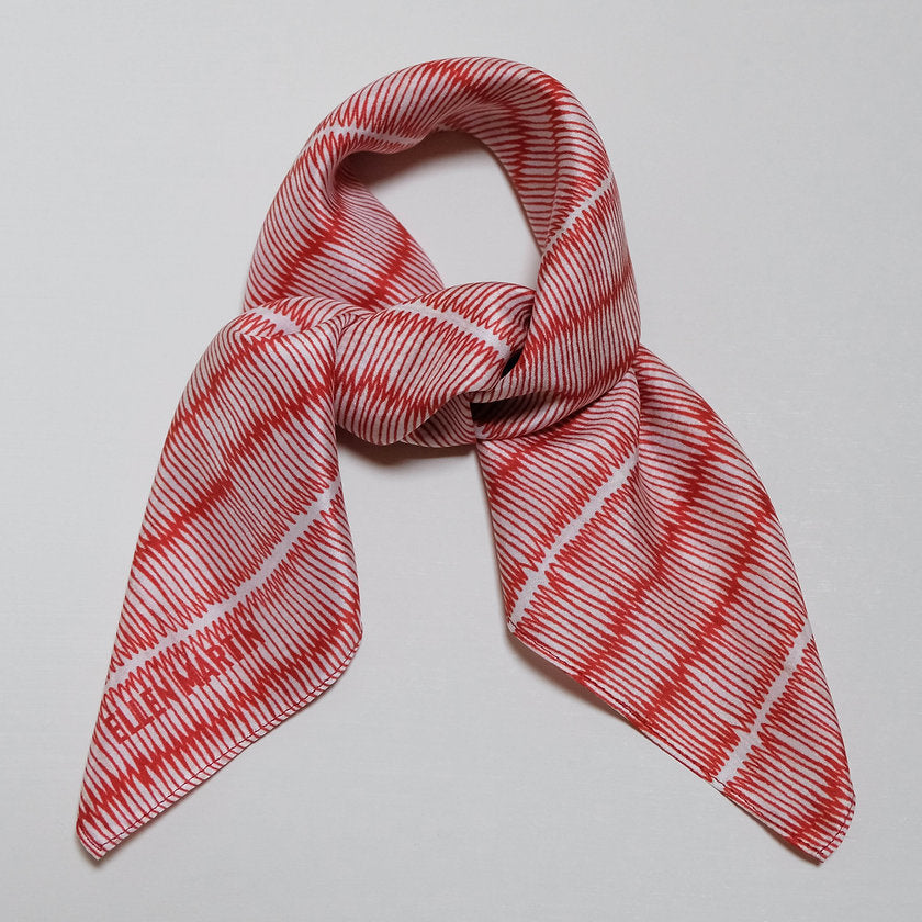Small square silk scarf in red and cream in an intricate repeat pattern, shown tied in bow against plain background.